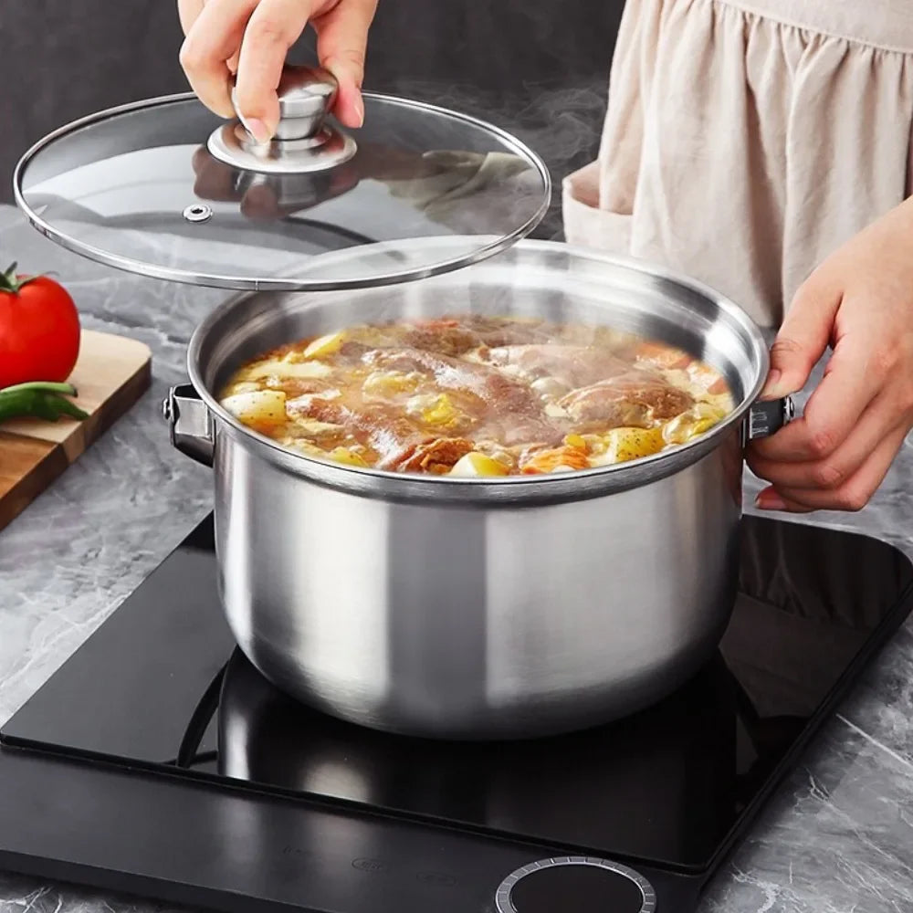 A stainless steel pot with a clear glass lid sits on a black induction cooktop, with a person lifting the lid to reveal simmering food inside. A tomato and green pepper are visible on a cutting board to the left, and the person wearing a beige apron is partially visible on the right.
