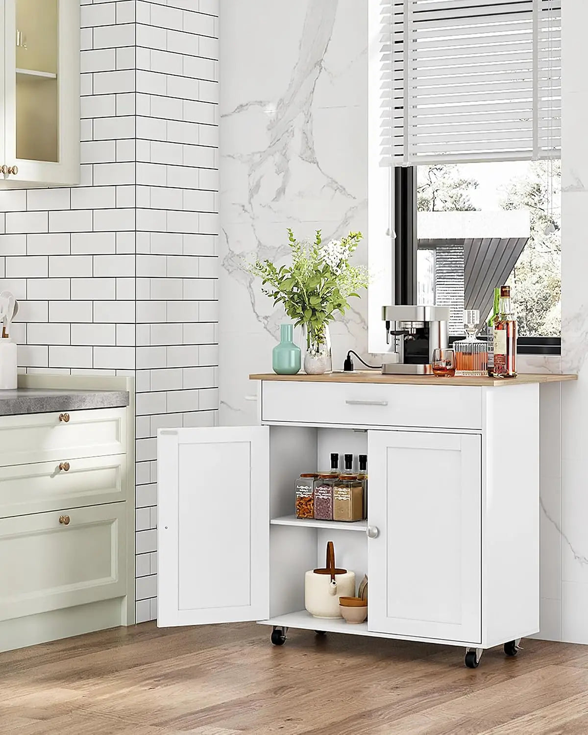 A white rolling kitchen cart with a light wood top stands in a bright kitchen. The cart has two front doors, one open to show shelves with jars and a small container, and is mounted on black caster wheels.