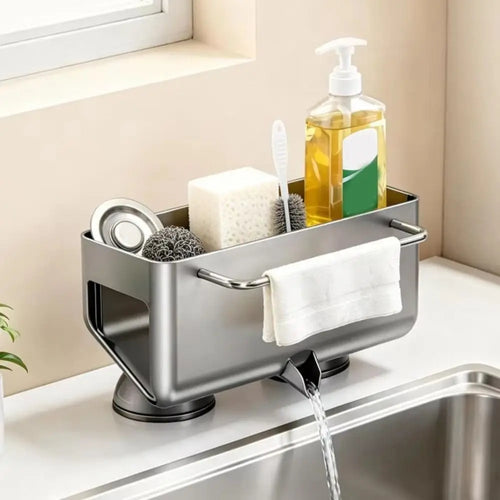 A stainless steel kitchen sink drain organizer with suction cups, shown holding cleaning supplies like a sponge, brush, soap dispenser, and a folded white towel. Water is flowing from the drain spout into the sink.