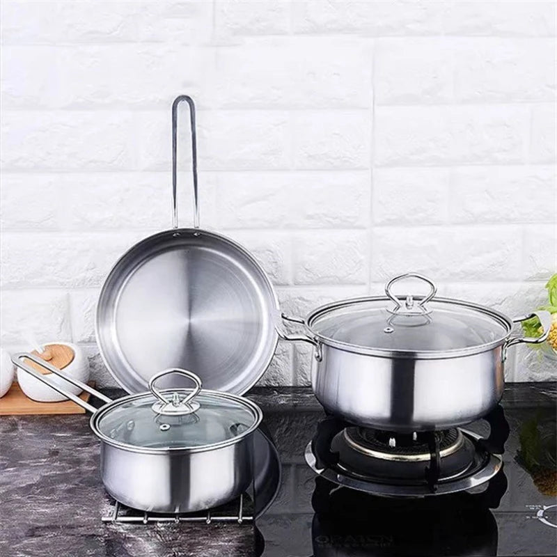 A stainless steel cookware set on a dark countertop against a white brick wall, including a large pot on a burner, a smaller pot with lid, and a frying pan leaning against the wall.