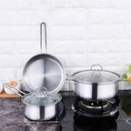 A stainless steel cookware set on a dark countertop against a white brick wall, including a large pot on a burner, a smaller pot with lid, and a frying pan leaning against the wall.