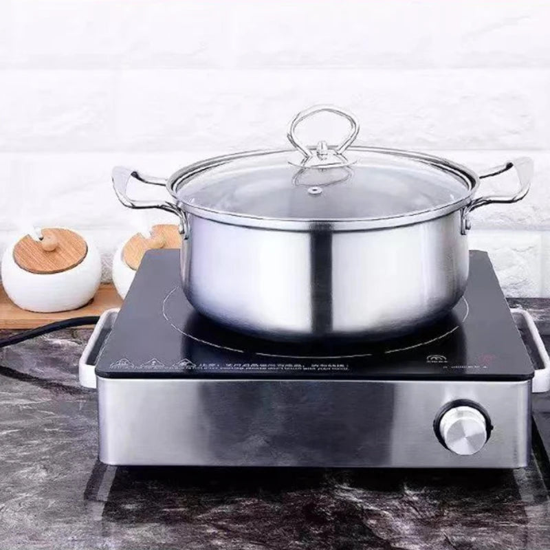 A stainless steel stockpot with a glass lid sits on a black and silver portable induction cooktop, set on a dark marble countertop against a white brick wall background.