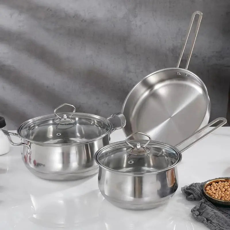 Three stainless steel cookware pieces on a white surface against a gray textured background: a small saucepan with lid in front, a medium pot with lid behind it, and a frying pan leaning against the back. A small bowl of peanuts and a gray cloth are partially visible at the lower right.