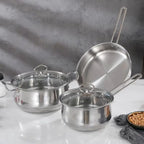 Three stainless steel cookware pieces on a white surface against a gray textured background: a small saucepan with lid in front, a medium pot with lid behind it, and a frying pan leaning against the back. A small bowl of peanuts and a gray cloth are partially visible at the lower right.