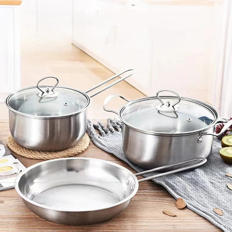 Three stainless steel cookware pieces on a wooden table in a bright kitchen setting: a small saucepan with glass lid, a large stockpot with glass lid, and a frying pan with long handle.