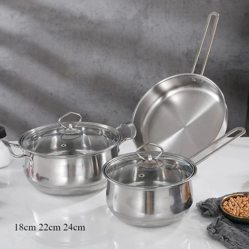 Three stainless steel cookware pieces with glass lids on a white countertop against a gray textured wall. A frying pan leans against the back, and two saucepans sit in front.