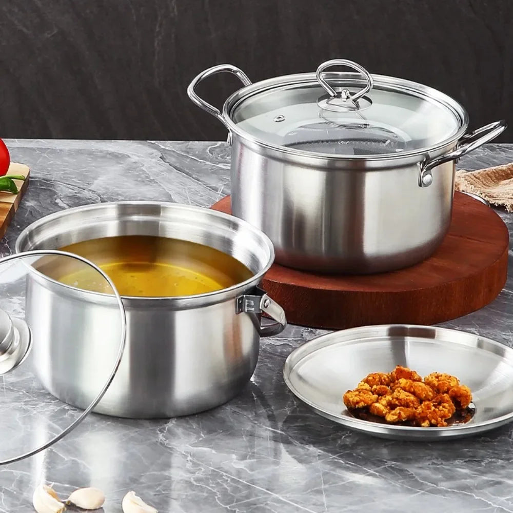 Two stainless steel stock pots with glass lids are shown on a gray marble countertop, one filled with yellow broth and one empty, with a small plate of fried food in front. A dark textured wall is in the background.
